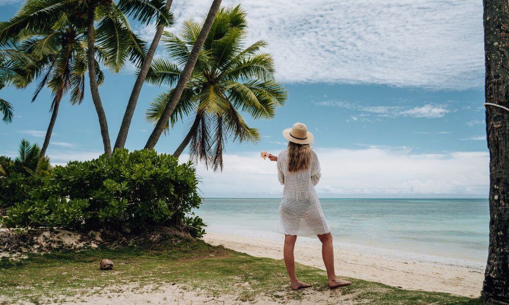Woman with a white dress and sunhat standing looking at a beach