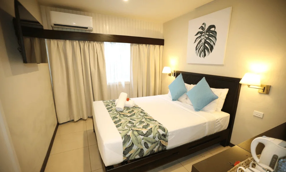 Small room with queen bed, blue accent pillows, and monstera leaf artwork at Fiji Gateway Hotel